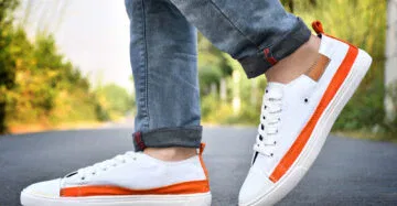 A pair of white and orange men's sneakers styled with rolled-up denim jeans, displayed on a road with a blurred natural background.