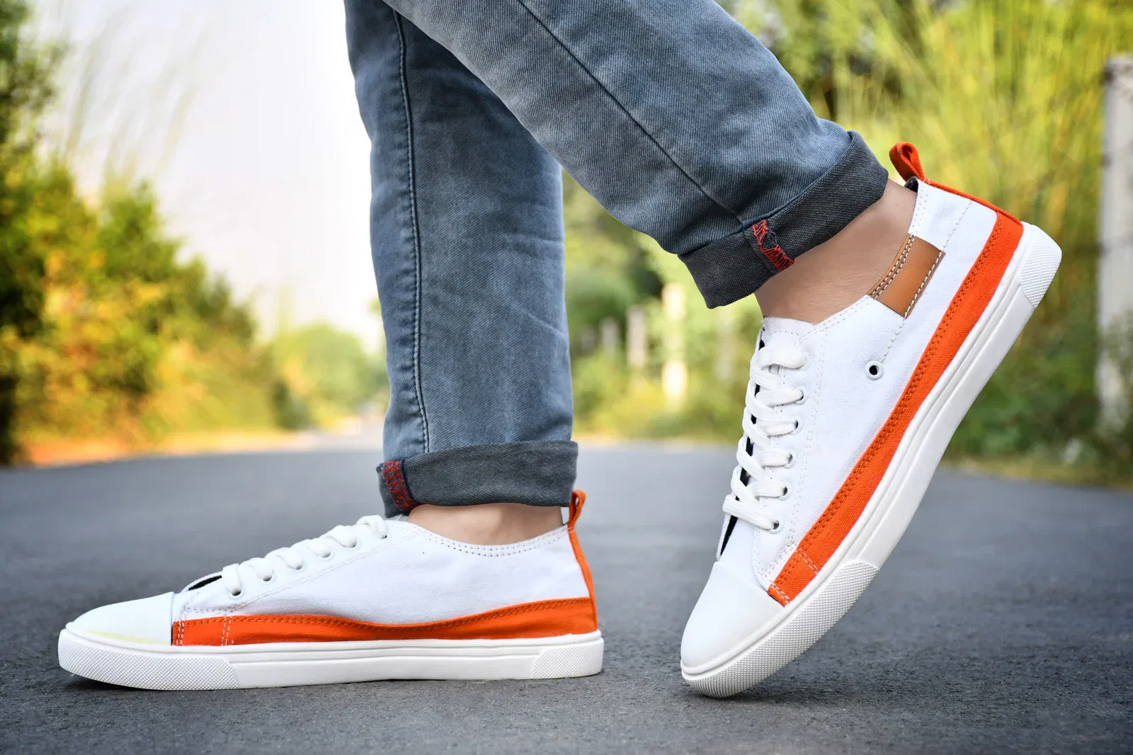 A pair of white and orange men's sneakers styled with rolled-up denim jeans, displayed on a road with a blurred natural background.